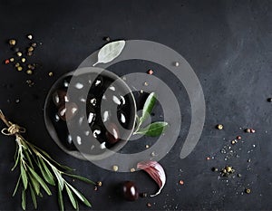 A bowl of delicious black olives, surrounded by herbs and spices, on a black stone table.