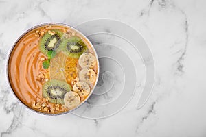 Bowl of delicious fruit smoothie with fresh banana, kiwi slices and granola on white marble table, top view. Space for text