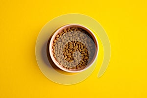 Bowl of dry kibble cat food on yellow background top view. Pet care concept