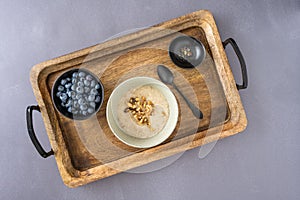 Bowl of fresh blueberries, small bowl of walnuts, and bowl of cooked oatmeal with spoon on a rustic wooden serving tray