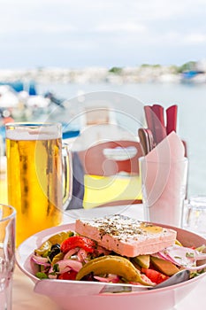 Bowl of fresh Greek salad with feta cheese. Greek salad and beer mug on table against the sea bay background in Greece