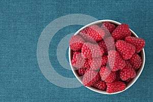 Bowl of fresh red raspberries for dessert, round white ceramic bowl, blue fabric background