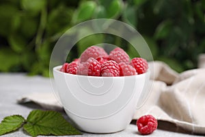 Bowl of fresh ripe raspberries with green leaves on light table against blurred background