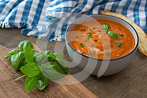 A bowl of fresh tomato soup in white ceramic bowl, garnished with basil, croutons, seasoning and a drizzle of olive oil