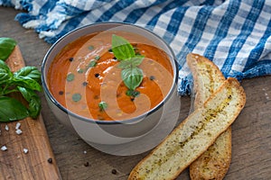 A bowl of fresh tomato soup in white ceramic bowl, garnished with basil, croutons, seasoning and a drizzle of olive oil