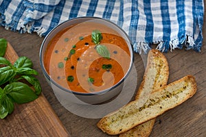 A bowl of fresh tomato soup in white ceramic bowl, garnished with basil, croutons, seasoning and a drizzle of olive oil