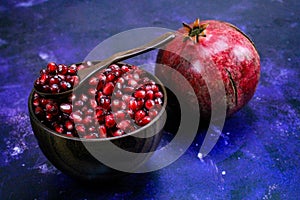 A bowl full of pomegranate seeds with a wooden spoon on it. A whole ripe pomegranate next to the bowl. Seeds and pomegranate are