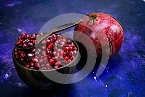 A bowl full of pomegranate seeds with a wooden spoon on it. A whole ripe pomegranate next to the bowl. Seeds and pomegranate are