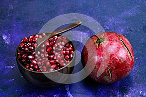 A bowl full of pomegranate seeds with a wooden spoon on it. A whole ripe pomegranate next to the bowl. Seeds and pomegranate are