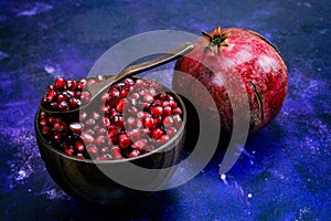 A bowl full of pomegranate seeds with a wooden spoon on it. A whole ripe pomegranate next to the bowl. Seeds and pomegranate are