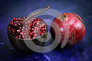 A bowl full of pomegranate seeds with a wooden spoon on it. A whole ripe pomegranate next to the bowl. Seeds and pomegranate are