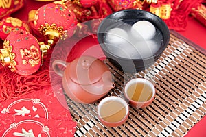 A bowl of glutinous rice balls, tea and red envelopes on the background of festivity. The Chinese characters in the picture mean `