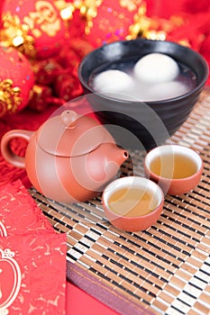 A bowl of glutinous rice balls, tea and red envelopes on the background of festivity. The Chinese characters in the picture mean `