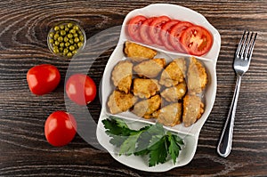 Bowl with green peas, tomatoes, partitioned dish with patties, parsley, slices of tomato, fork on table. Top view