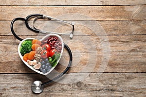 Bowl with heart-healthy diet products and stethoscope on wooden background, top view