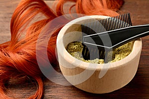 Bowl of henna powder, brush and red strand on wooden table, closeup. Natural hair coloring