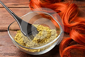 Bowl of henna powder, brush and red strand on wooden table, closeup. Natural hair coloring