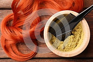 Bowl of henna powder, brush and red strand on wooden table, flat lay. Natural hair coloring