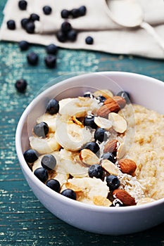Bowl of homemade oatmeal porridge with banana, blueberries, almonds, coconut and caramel sauce on teal rustic table