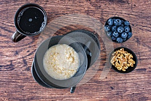 Bowl of hot oatmeal, black bowl and plate, black spoon, small bowls of blueberries and walnuts, wood table, cup of tea