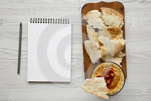 Bowl of Hummus with chickpeas, paprika, olive oil and pita bread on rustic wooden board. Blank notepad with pencil. White wooden s