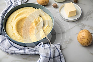 Bowl with mashed potatoes and ingredients as potato, milk, salt, butter, nutmeg with kitchen towel and grinder on light marble