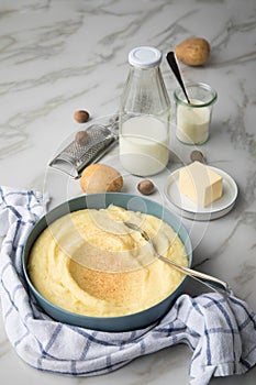 Bowl with mashed potatoes and ingredients as potato, milk, salt, butter, nutmeg with kitchen towel and grinder on light marble