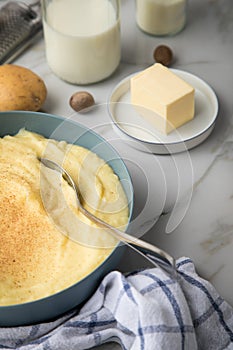 Bowl with mashed potatoes and ingredients as potato, milk, salt, butter, nutmeg with kitchen towel and grinder on light marble