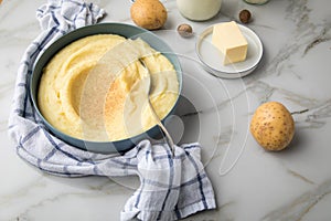 Bowl with mashed potatoes and ingredients as potato, milk, salt, butter, nutmeg with kitchen towel and grinder on light marble