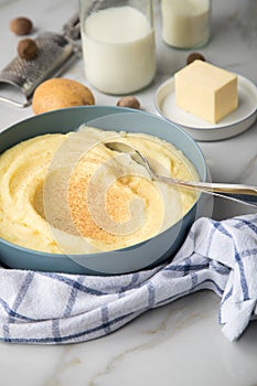 Bowl with mashed potatoes and ingredients as potato, milk, salt, butter, nutmeg with kitchen towel and grinder on light marble