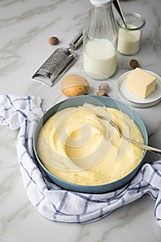 Bowl with mashed potatoes and ingredients as potato, milk, salt, butter, nutmeg with kitchen towel and grinder on light marble