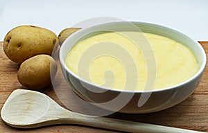 A bowl of mashed potatoes puree, with raw potatoes and wooden spoon.