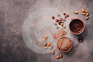 A bowl of melted chocolate, cocoa powder, almonds and hazelnuts on a dark rustic background. Top view, flat lay, copy space