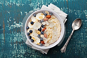 Bowl of oatmeal porridge with banana, blueberries, almonds, coconut and caramel sauce on teal rustic table, top view, flat lay