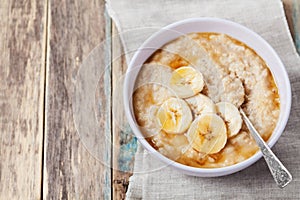 Bowl of oatmeal porridge with banana and caramel sauce on rustic table, hot and healthy breakfast