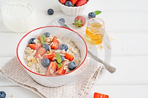 Bowl of oatmeal porridge with blueberries, strawberries, almond petals and honey on a white wooden background.