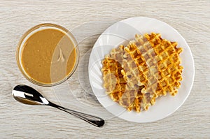 Bowl with peanut butter, spoon, bowl with waffle on wooden table. Top view
