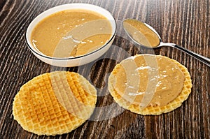 Bowl with peanut paste, sandwich with peanut butter, wafer, spoon with peanut paste on table