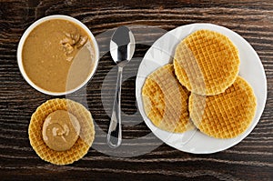 Bowl with peanut paste, spoon, sandwich with peanut butter, wafers in plate on wooden table. Top view