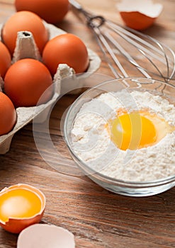 Bowl plate with flour and egg yolk on wooden background with fresh raw eggs and metal whisk