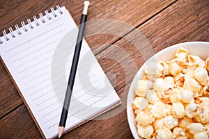 A bowl of popcorn and notebook and Black pencil on a wooden table.