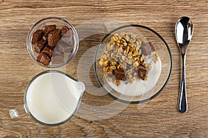 Bowl with porous chocolate, pitcher with yogurt, bowl with granola, yogurt, chocolate, spoon on table. Top view