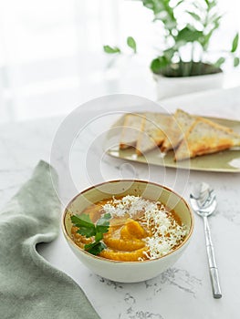 Bowl of pumpkin or carrot vegetarian cream soup decorated by fresh parsley and grated cheddar cheese white marble table