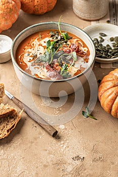 Bowl of pumpkin cream soup decorated with cream, pumpkin seeds, bacon, microgreens and olive oil on brown textured