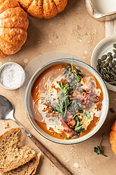 Bowl of pumpkin cream soup decorated with pumpkin seeds, bacon, microgreens and olive oil on brown textured background