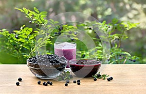 Bowl with ripe berries wild bilberries, bowl with jam of bilberries and of fruit yogurt on wooden table on background of shrubs