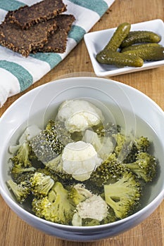 Bowl of soup with pieces of chicken, broccoli and cauliflower, bread and cucumbers on towel