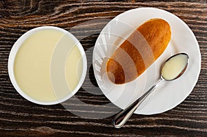 Bowl with sweet condensed milk, bun and spoon with condensed milk in plate on wooden table. Top view