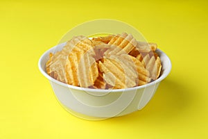 Bowl of tasty ridged potato chips on yellow background