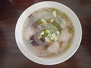 a bowl of tekwan soup in a white bowl topped with sliced ??cucumber, celery leaves, fried onions and ear mushrooms